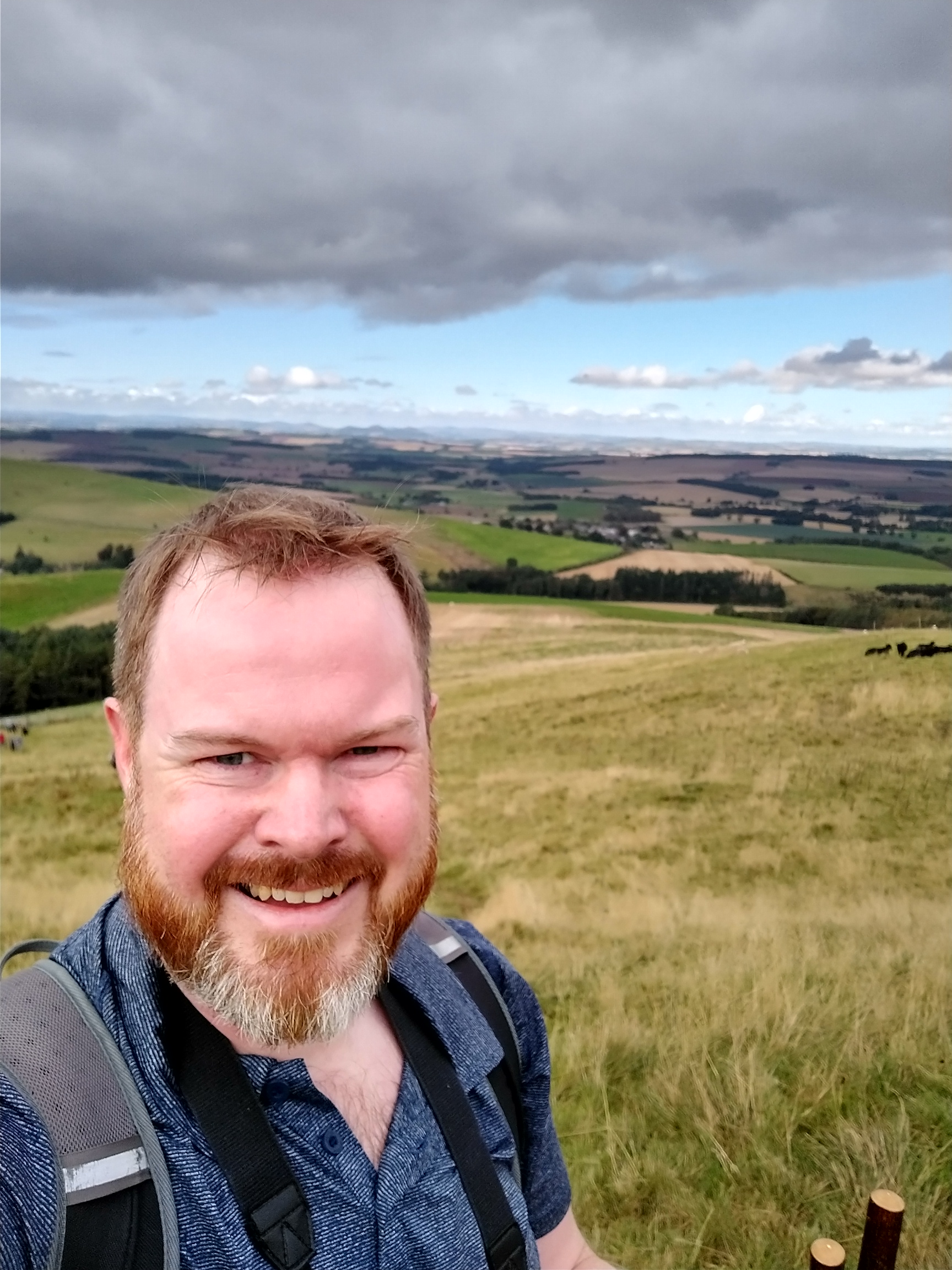 A selfie with Mark smiling and windswept in the foreground, a wide grassy hillside behind him. A cloudy grey sky, with blue in the distance.